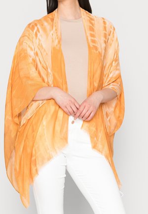 Frau trägt einen orange gemusterten, transparenten Schal über einem beigen Oberteil und weißen Hosen, mit verschränkten Händen vor dem Körper.