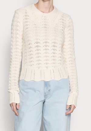 Femme portant un pull en tricot couleur crème avec un col et un ourlet festonnés, assorti à un jean taille haute bleu clair.