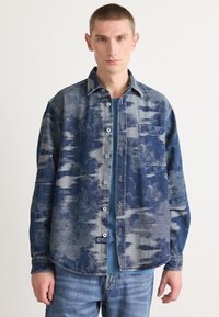 Blauwe denimschoen met een textuurpatroon, knopen aan de voorkant en een borstzak. Draag het over een blauwe t-shirt. Casual fit met lange mouwen.