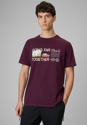 Młody mężczyzna w bordowej koszulce z grafiką samochodu i przyczepy kempingowej oraz napisem „Going Far Together” na jednolitym szarym tle.