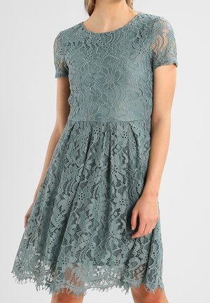 Kobieta ubrana w koronkową sukienkę do kolan o krótkich rękawach w kolorze morsko-zielonym, z kwiatowym wzorem i falowanym brzegiem.