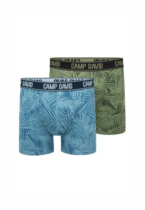Oberkörperfreier Mann trägt blaue gemusterte Boxerbriefs mit "CAMP DAVID" Bund, steht vor einem einfarbigen hellen Hintergrund.