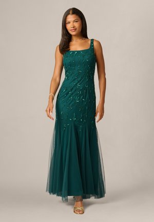 Vrouw draagt een lange turquoise jurk met bloemrijke kralenborduursel en een doorschijnende overlay, staand tegen een effen neutrale achtergrond, draagt gouden sandalen.