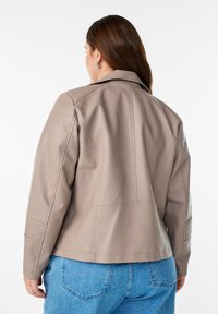 Beige Kunstlederjacke mit strukturiertem Design, gepolsterten Schultern und Nähten, kombiniert mit hellblauen Jeans.