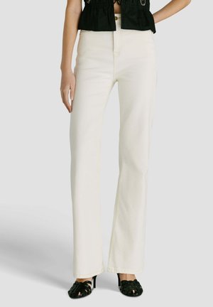 Vrouw draagt hoge taille, rechte pijpen witte broek gecombineerd met zwarte sandalen met hak en gevlochten bandjes en een cropped zwart topje.