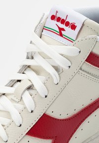 High-top sneaker in white leather with red accents, featuring white laces, a padded collar, and a woven logo label at the top.