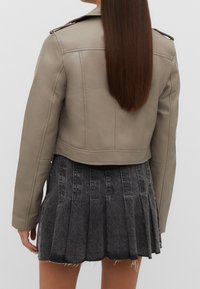 Beige cropped faux leather jacket with shoulder epaulettes, paired with a black denim pleated skirt featuring raw hem.