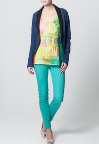 Blå blazer över en flerfärgad linne med text, kombinerad med turkosa jeans och metalliska högklackade skor. Släta tyger, livfulla färger.