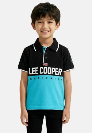 Garçon souriant portant un polo noir et bleu avec le texte "Lee Cooper Authentic" et un petit drapeau Union Jack sur la poitrine, debout devant un fond blanc.