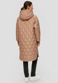 Vrouw in een lange, gewatteerde beige jas met capuchon, zwarte broek en witte sneakers, staand in zijaanzicht op een lichtgrijze achtergrond.
