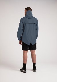 Chaqueta impermeable en azul-gris con capucha, mangas largas y un dobladillo trasero más largo, combinada con pantalones cortos negros y zapatillas deportivas.