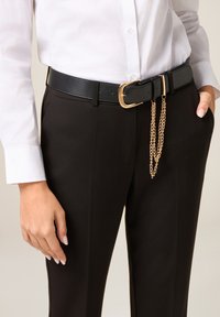 Cinturón de cuero negro con hebilla rectangular de tono dorado y detalle decorativo de cadena, llevado sobre pantalones negros y una camisa blanca.