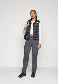 Czarna puchowa kamizelka, szare wodoodporne spodnie i czarne trampki. Biała koszula z długim rękawem i skróconym dołem, minimalistyczny design, funkcjonalny styl.