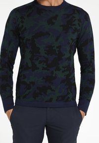 Homme portant un pull à manches longues à motif camouflage bleu marine et vert avec un pantalon bleu marine, les mains dans les poches.