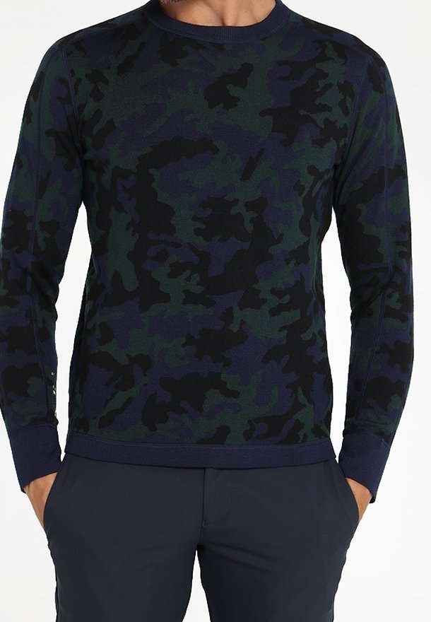 Homme portant un pull à manches longues à motif camouflage bleu marine et vert avec un pantalon bleu marine, les mains dans les poches.