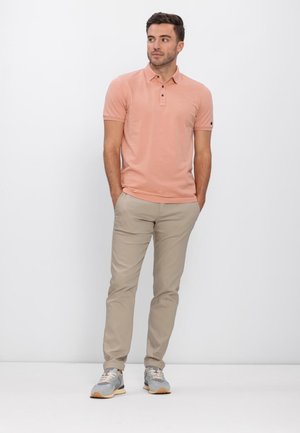 Homme debout les mains dans les poches, portant un polo rose saumon, un pantalon beige et des baskets grises, sur un fond blanc.