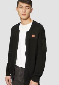 Zwarte zip-up cardigan met geribbelde textuur, voorzien van een bruin leren logo label en zijzakdetails. Draag over een wit T-shirt.