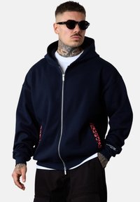 Homme avec des tatouages sur le cou portant des lunettes noires, un sweat à capuche zippé bleu marine avec des accents de poche rouges à motifs, un t-shirt blanc et un pantalon noir.