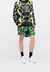 Personne portant une chemise à manches longues à motif abstrait vert et noir avec un short assorti vert et noir, des chaussettes noires et des baskets blanches.