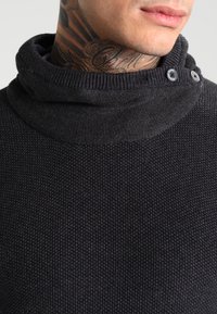 Pull noir en maille avec col entonnoir, présentant un tissu texturé, des poignets côtelés et des détails de boutons sur un côté du col.