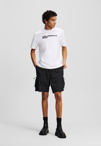 Weiße T-Shirt mit kurzen Ärmeln und dem "KARL LAGERFELD JEANS" Logo, kombiniert mit schwarzen Cargo-Shorts, die mehrere Taschen und Kordeln haben.