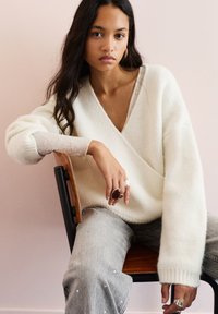 Luźny biały sweter z dekoltem w serek, teksturowane rękawy; szare dżinsy z perłowymi zdobieniami. Osoba siedząca na drewnianym krześle.