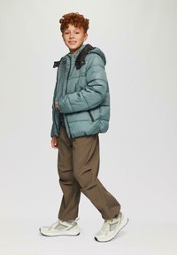 Gevoerde jas in teal met zwarte accenten, lichtgewicht materiaal, met ritszakken, gedragen over een grijze trui, gecombineerd met een bruine broek en sneakers.
