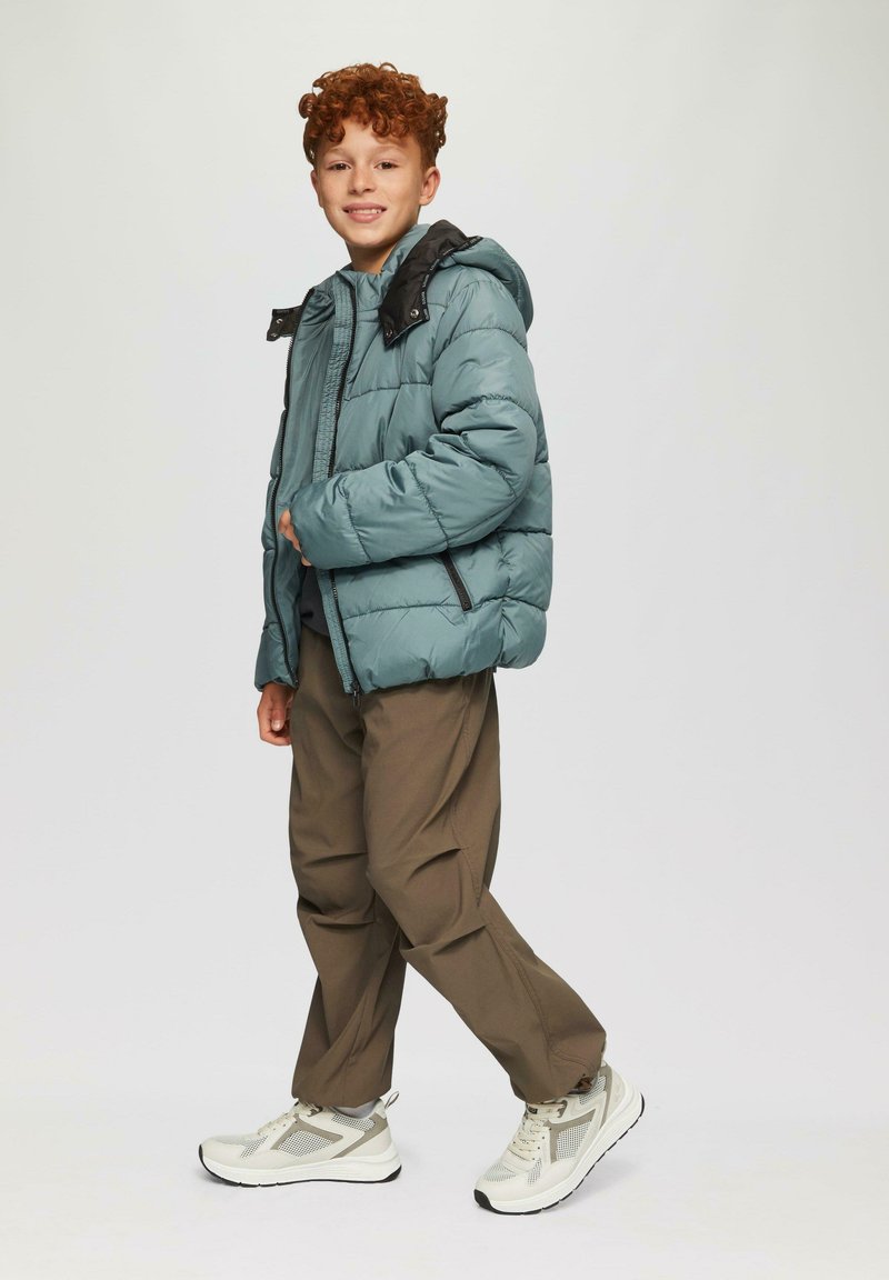 Gevoerde jas in teal met zwarte accenten, lichtgewicht materiaal, met ritszakken, gedragen over een grijze trui, gecombineerd met een bruine broek en sneakers.