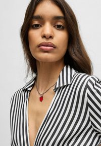 Gestreepte zwart-witte shirt met een diepe V-hals, voorzien van een zilveren ketting met een rode hartpendant met "HUGO" gegraveerd.