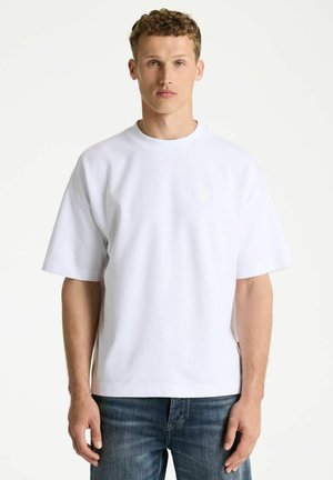 Young man with curly hair wearing a plain white oversized short-sleeve shirt and blue jeans, facing forward against a white background.