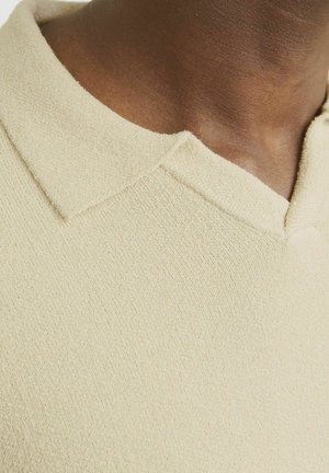 Hellbeiges Poloshirt mit strukturiertem Stoff und flachem Kragen. Der Halsausschnitt hat eine leichte Öffnung, die eine glatte Textur zeigt.