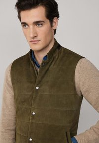Olivově zelená semišová vesta s vysokým límcem, zapínáním na knoflíky a bočními kapsami. Má texturovaný vzor a kontrastní černé akcenty na ramenou.
