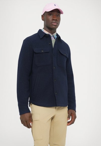 Casaco camisa azul-marinho de mistura de lã com dois bolsos no peito e bolsos laterais, fecho de botões e textura suave, usado sobre uma camisa de padrão xadrez claro.