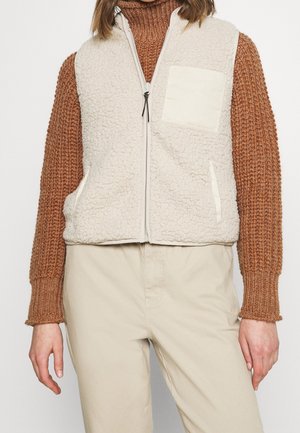 Personen bærer cremefarvet fleecevest med lynlås over brun strikket sweater og beige bukser, stående mod en ensfarvet hvid baggrund.