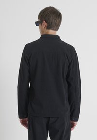 Camisa negra de manga larga hecha de tela texturizada, con cuello y puños abotonados. La parte posterior es lisa con un dobladillo recto.