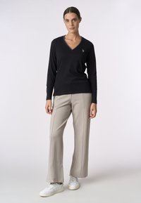 Maglietta nera a maniche lunghe con scollo a V e un piccolo logo sul petto, abbinata a pantaloni beige a gamba larga e scarpe da ginnastica bianche. Tessuto morbido.