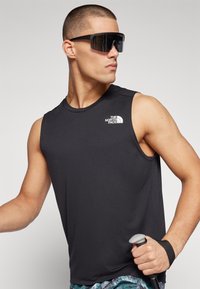 Μαύρο αμάνικο αθλητικό μπλουζάκι με λογότυπο στο στήθος, εφαρμοστό σχήμα, λείος ύφασμα, συνδυασμένο με μεγάλα γυαλιά ηλίου και κρατώντας ένα μπαστούνι πεζοπορίας.