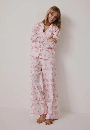 Vrouw in roze patroon lange mouwen pyjama staat blootsvoets tegen een lichte muur, glimlachend met gekruiste armen.
