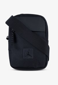 Black fabric sling bag with a textured surface, featuring a zippered pocket, adjustable strap, and a circular logo patch.