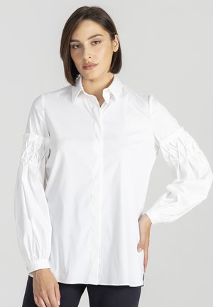 Kobieta z krótkimi brązowymi włosami, ubrana w białą koszulę z długim rękawem i guzikami, z plisowanymi detalami na rękawach, na jednolitym szarym tle.