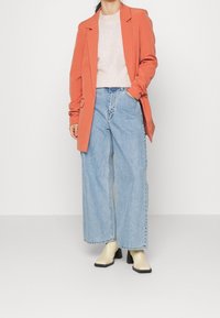 Blazer orange sur un pull gris clair, associé à un jean large bleu clair et des bottines crème à talons noirs.