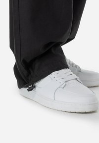 Black loose-fitting pants with a toggle closure at the hem, paired with white sneakers featuring textured detailing and perforations.