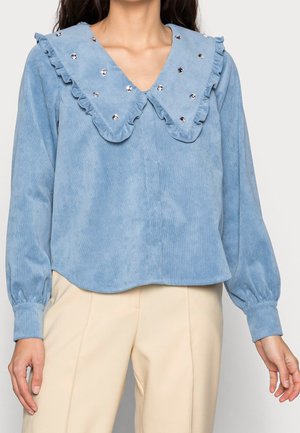 Lichtblauwe blouse met lange mouwen en oversized ruches met zilveren studs, gecombineerd met beige broek.