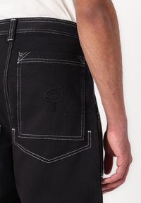 Sorte denimbukser har hvite sømmer, en baklomme med logo og unikt sømdesign, noe som fremhever en robust og funksjonell konstruksjon.