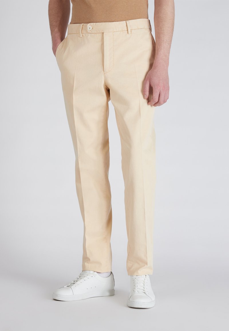 Uomo che indossa pantaloni beige a gamba dritta e sneakers bianche, in piedi con una mano in tasca contro uno sfondo neutro.