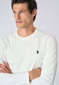 T-shirt a maniche lunghe beige chiaro realizzato in tessuto morbido, con un piccolo logo nero sul petto sinistro e un collo rotondo.