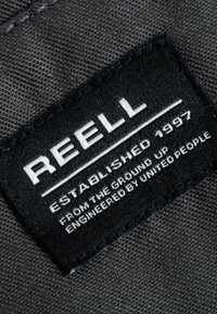 Black fabric label with white text stating "REELL," "ESTABLISHED 1997," and "ENGINEERED BY UNITED PEOPLE" on a textured gray background.