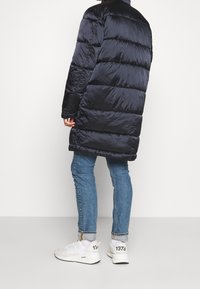 Abrigo acolchado azul marino con diseño acolchado, largo hasta los hombros, mangas largas y cuello alto. Se lleva con jeans azul claro y zapatillas blancas.