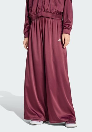 Weit geschnittene bordeauxrote Satinhose mit elastischem Bund und kleinem weißem Logo am linken Oberschenkel, getragen mit passender Jacke und lila Turnschuhen.