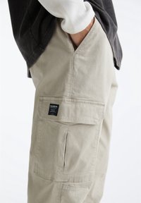 Beige cargo pants feature a side pocket, stitched detailing, and a label tag. Worn with a dark top and white long-sleeve shirt underneath.